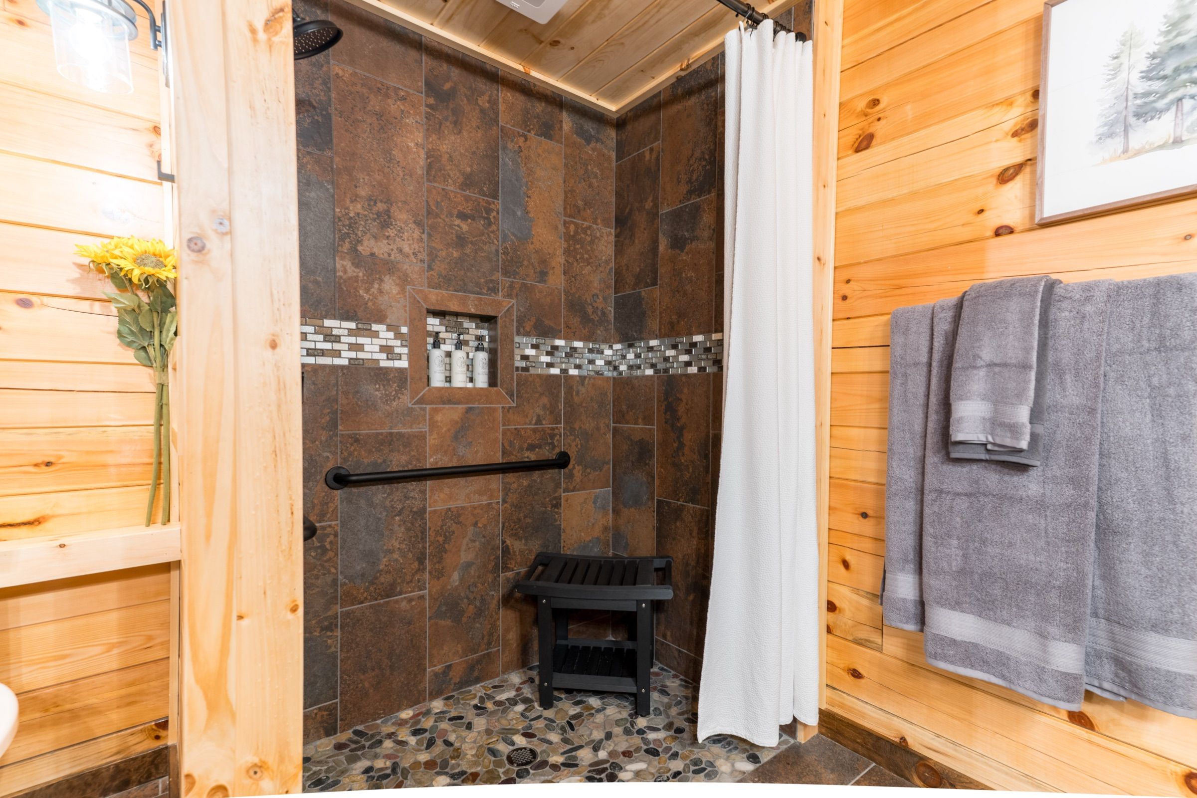 The roll-in shower: tiled walls, a pebble floor level with the bathroom floor, no curb, a black grab bar on the wall, a black shower bench, and a white curtain instead of a door.