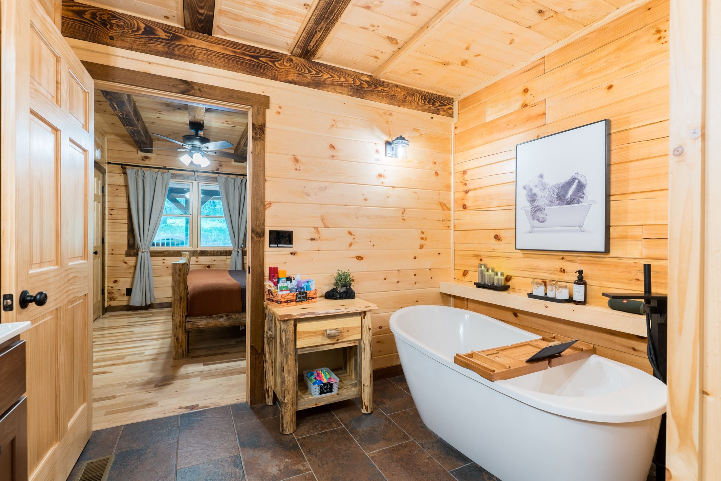 The accessible bathroom: a freestanding soaking tub, tile floor, and a wide open doorway through to the main-floor king suite with its log bed and window.
