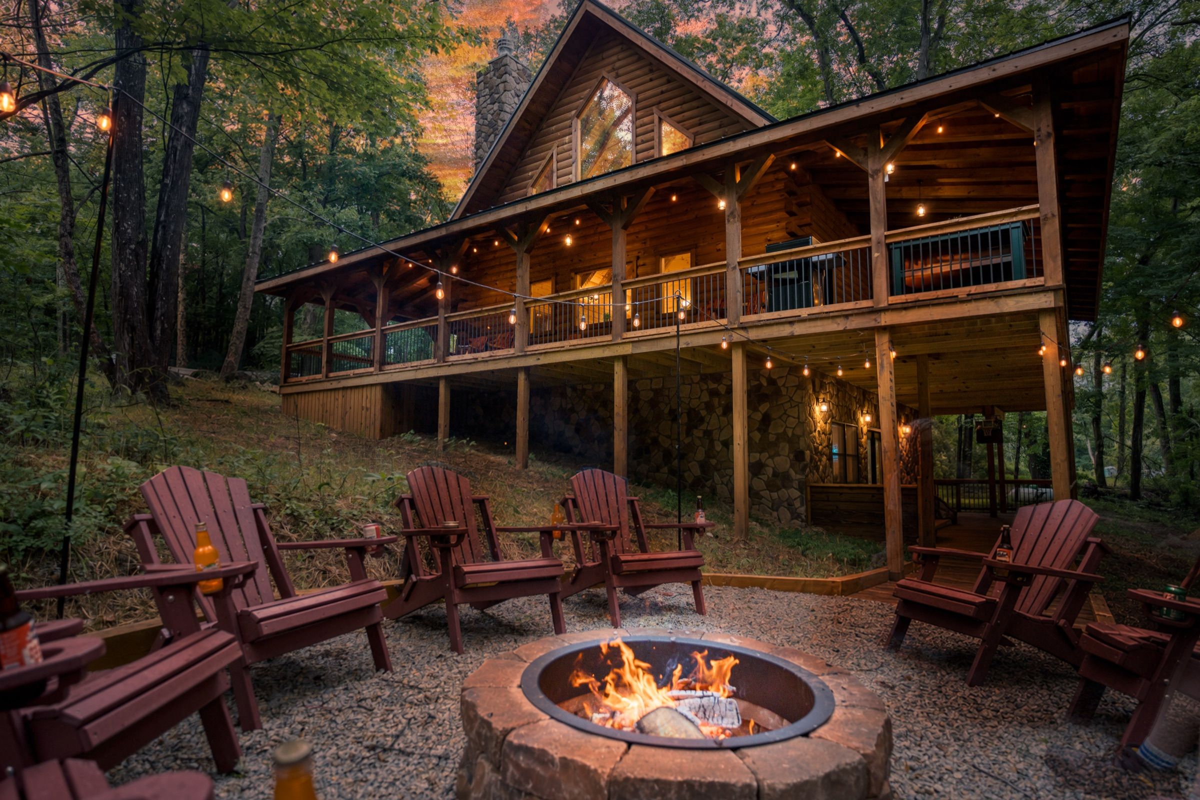The lodge at dusk from the fire pit: a ring of red Adirondack chairs around a stone fire ring with a fire burning, string lights across the deck, and warm light in every window under a pink sky.