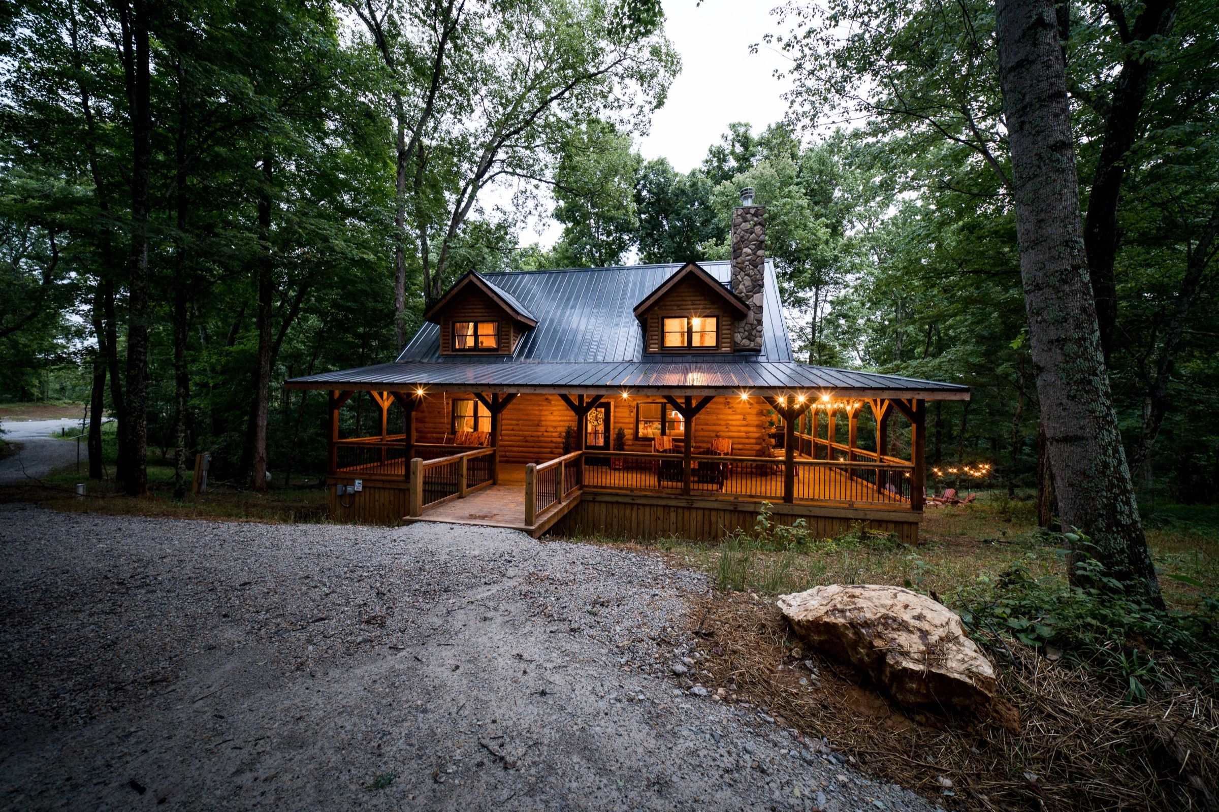Sleepy Bear Lodge from the gravel drive at dusk: a log cabin with a black metal roof, two dormers, a stone chimney, and a lit wrap-around porch.