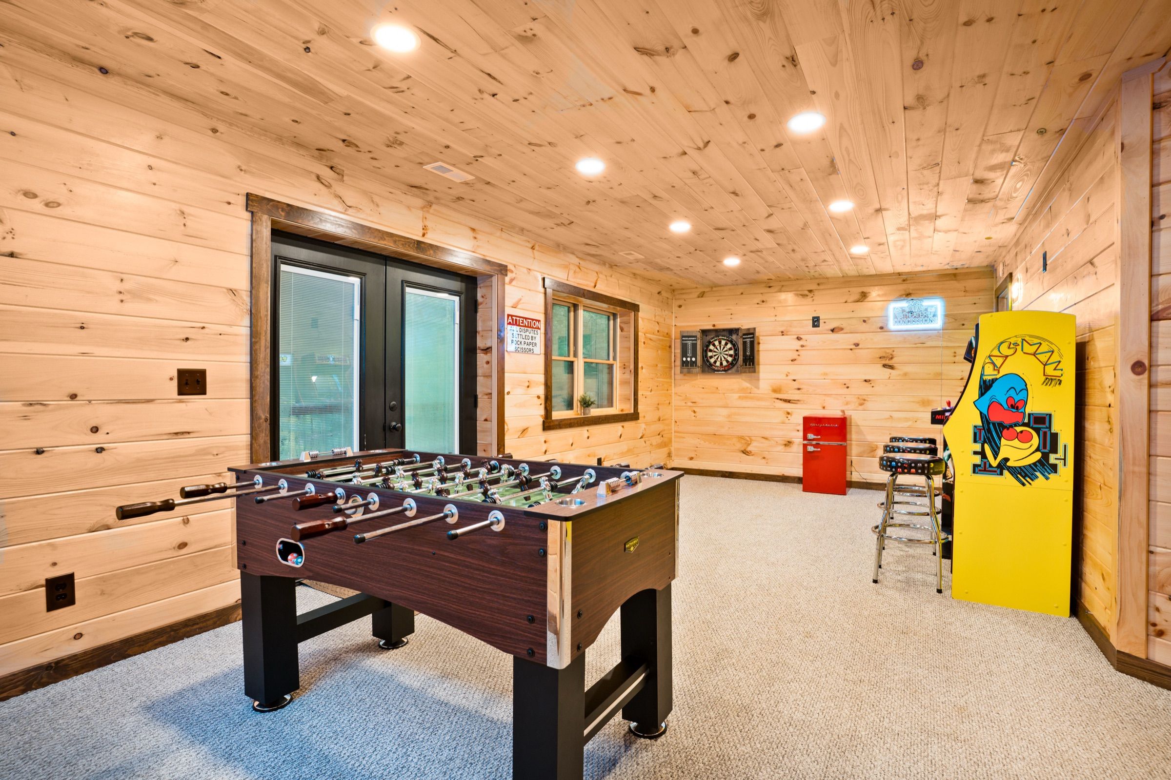 Game room with a foosball table, a yellow Pac-Man arcade cabinet, a dartboard, a red mini fridge, and bar stools.