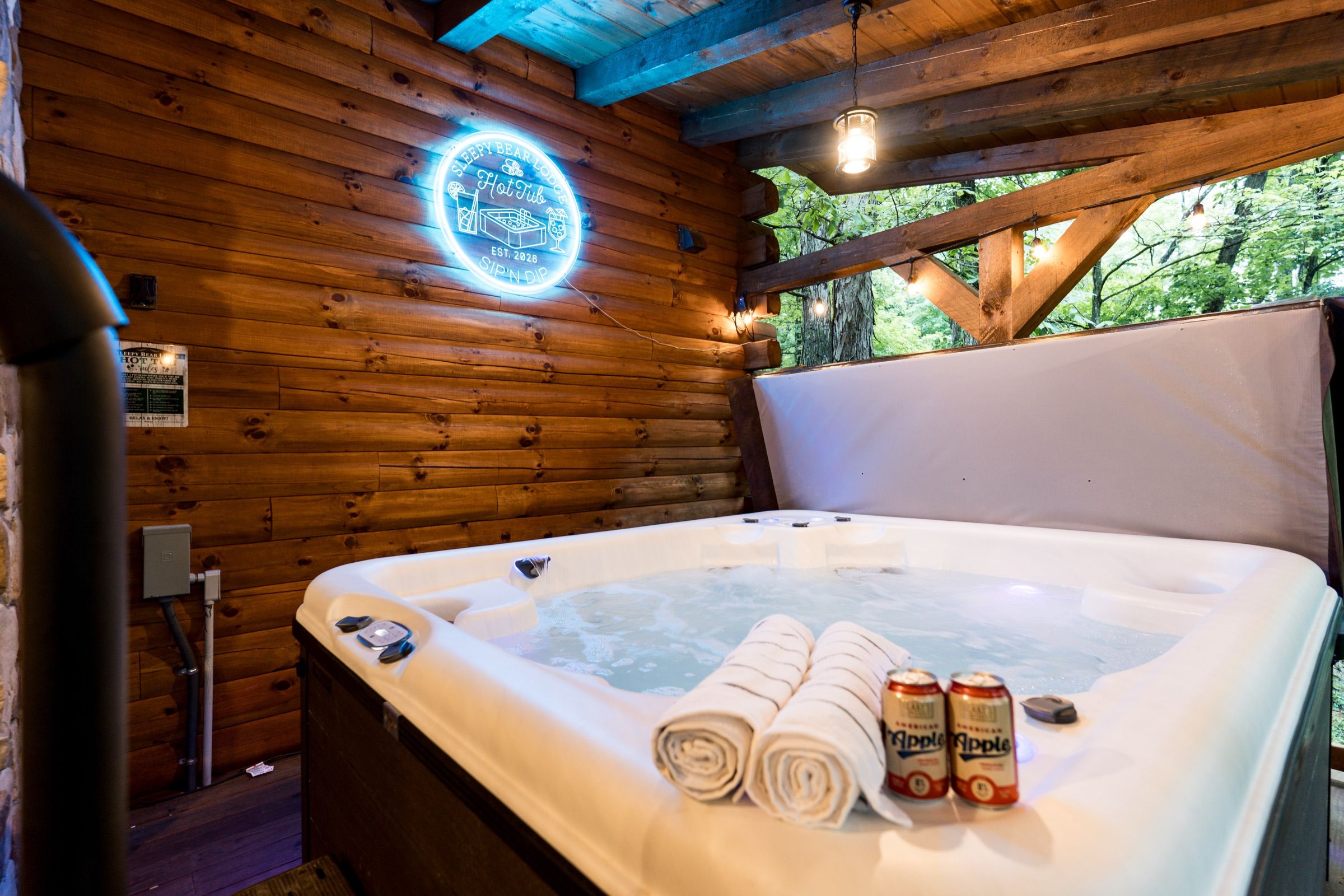 Hot tub on the covered deck at night, towels rolled on the rim, a blue neon sign reading Sleepy Bear Lodge Hot Tub, Sip 'n Dip on the log wall.