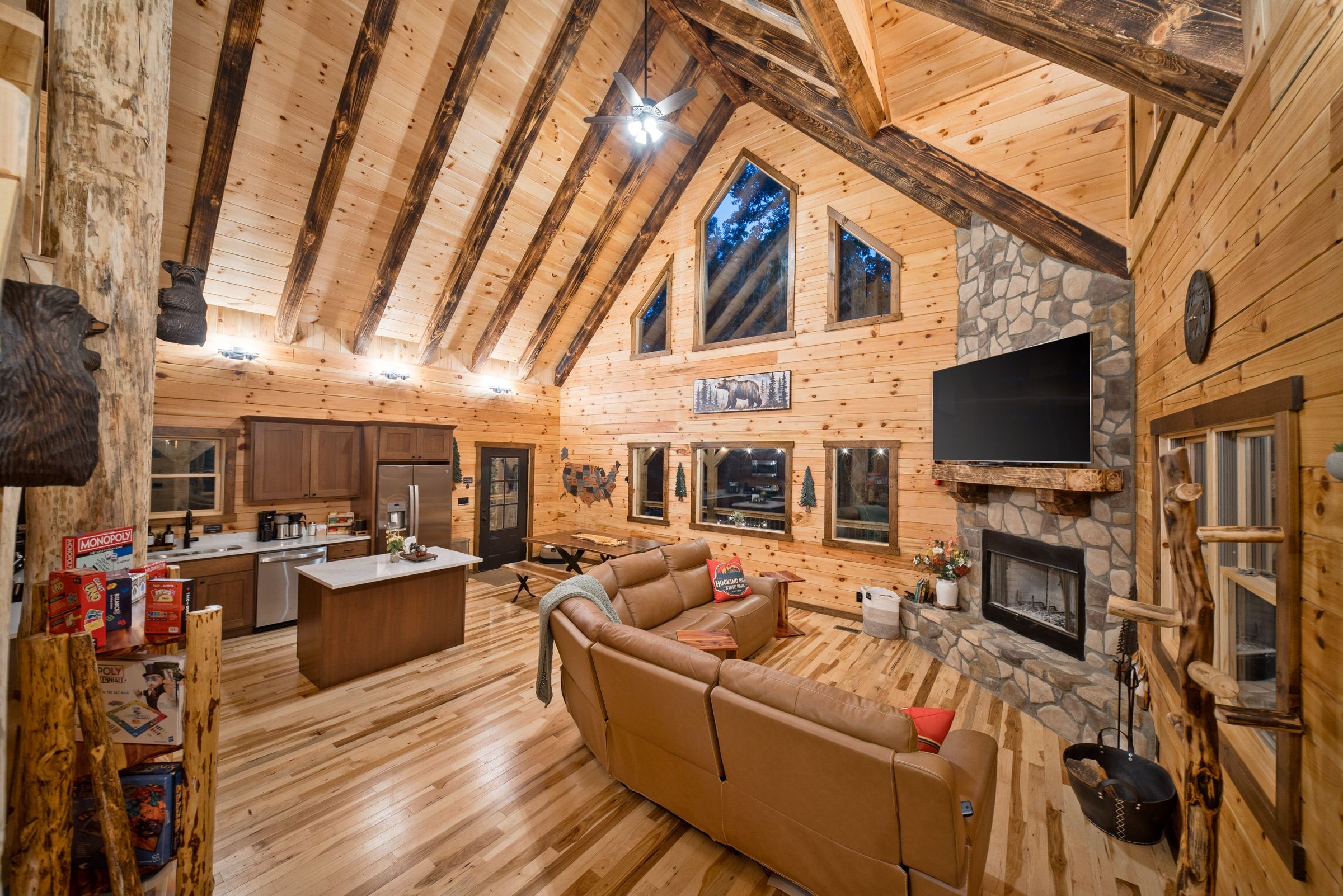 The great room at dusk: vaulted pine ceiling with charred beams, a stone fireplace with a mounted TV, a tan leather sectional, and the open kitchen behind it; blue evening light in the gable windows.